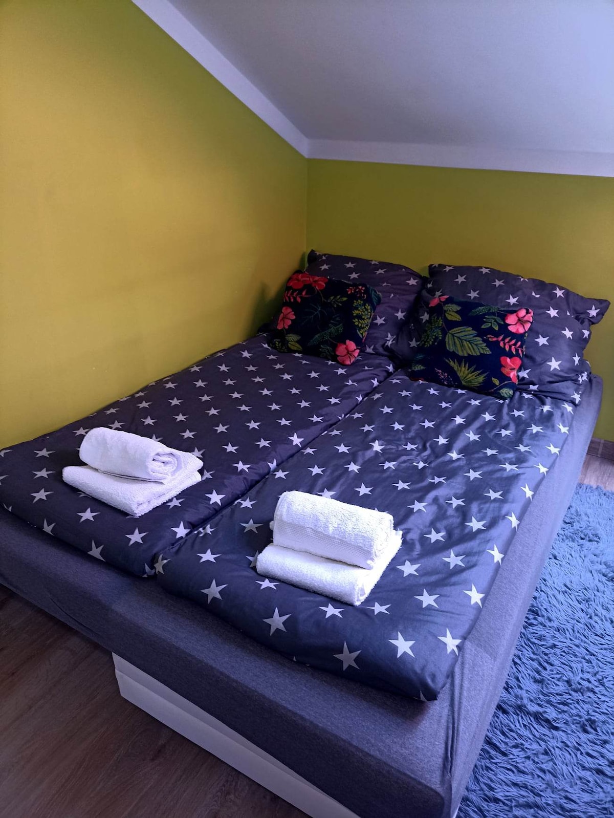 A sleeping area features a spacious bed dressed in star-patterned bedding with colorful floral pillows. Soft towels are neatly placed at the foot of the bed. The walls are painted in a calming green, enhancing the overall atmosphere of relaxation.