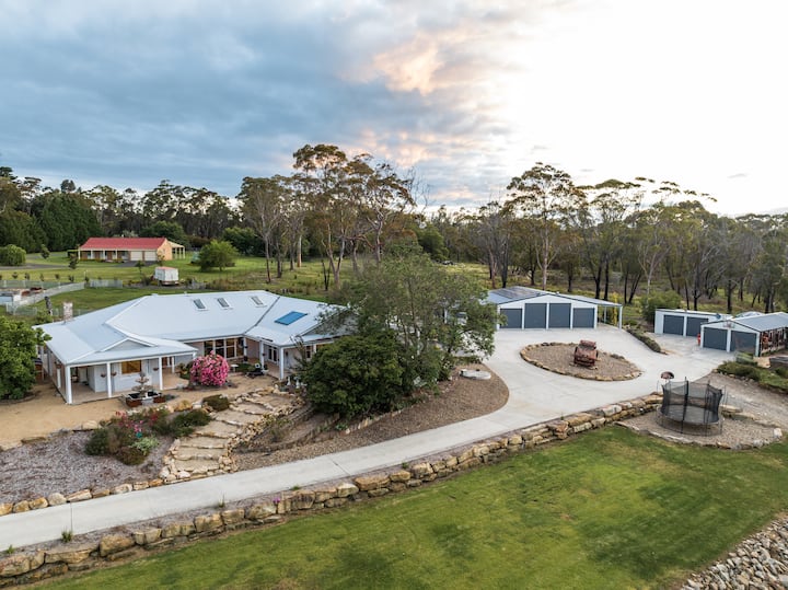 A modern house with a metal roof and large windows sits surrounded by trees and a spacious yard, with several sheds and a circular driveway leading to the main building. The sky is partly cloudy with evening light.