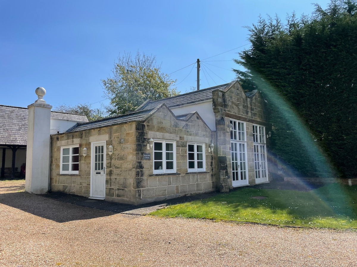 A charming exterior of a stone building is presented, featuring large windows that allow natural light to enter. Lush greenery surrounds the structure, while a gravel pathway leads to the entrance. The unique architectural details enhance the character of the property.