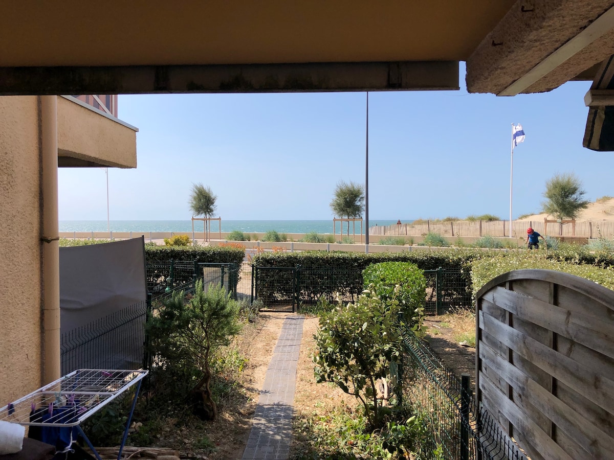 A view from the terrace shows a path leading through a garden towards the ocean. The sea is visible in the distance, framed by greenery along the sandy beach. Clear skies enhance the sense of openness in this coastal setting.