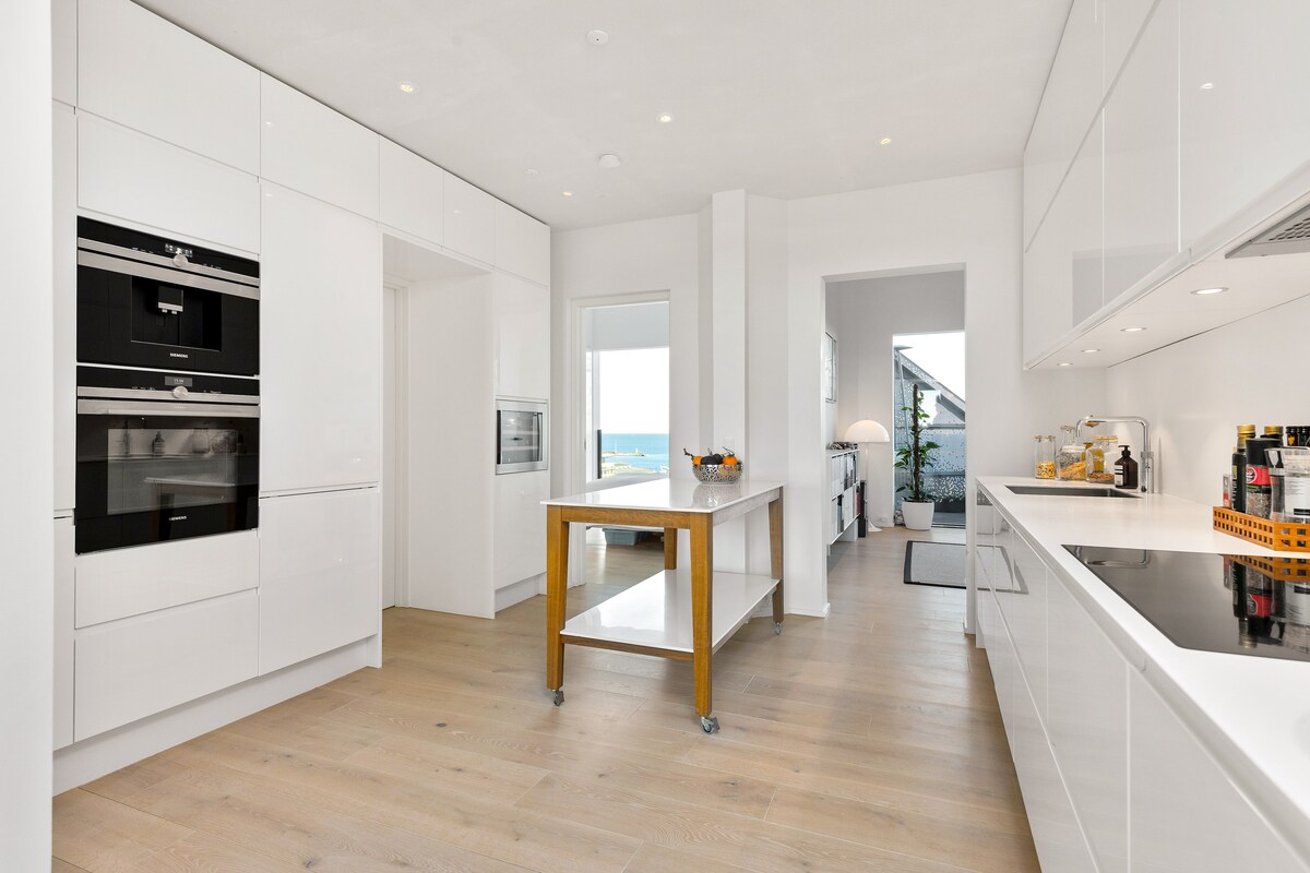 A modern kitchen is showcased with sleek, white cabinetry and high-end appliances. A central wooden island provides additional workspace. Natural light floods the area through large windows, highlighting the clean lines and spacious layout.