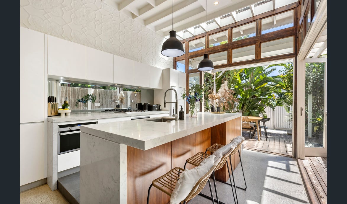 A modern kitchen features a large island with sleek countertops and stylish bar stools. Large windows allow natural light to fill the space, complemented by greenery visible outside. Ample cabinetry and contemporary appliances contribute to a functional design.