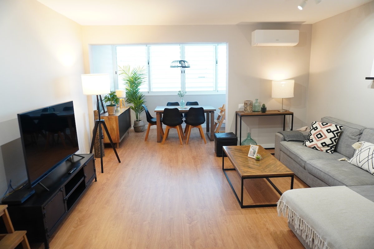 A bright living area features a comfortable gray sofa and a sleek coffee table made of wood. A dining space accommodates six chairs and a simple table, with a large window allowing natural light. Potted plants add greenery, enhancing the inviting ambiance.