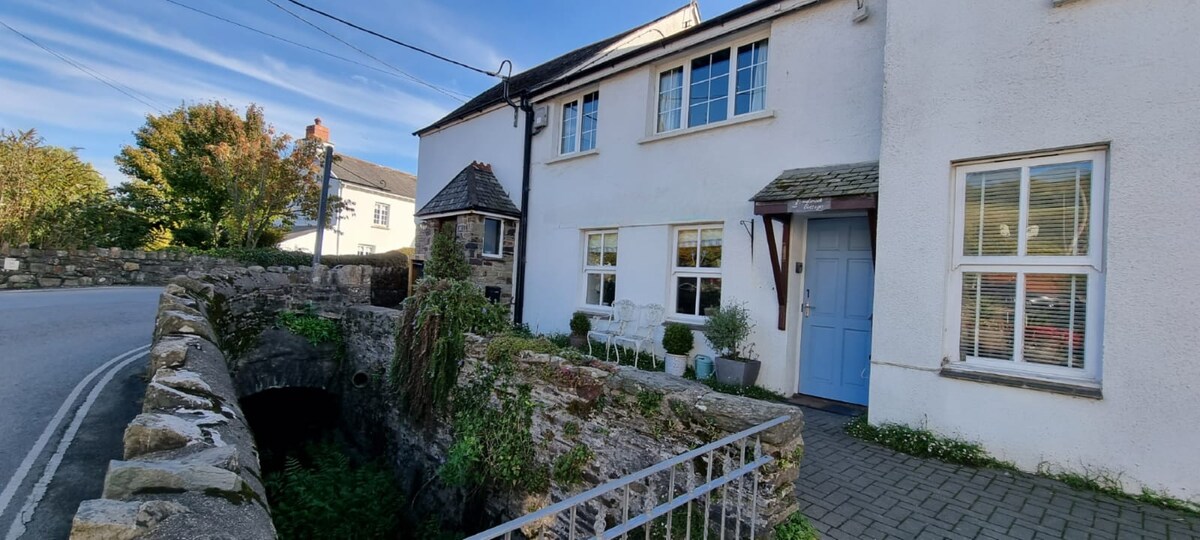 The cottage exterior showcases a charming white facade with blue door accents. A decorative stone wall surrounds a gentle stream, bordered by a paved pathway. Nearby greenery and a bench provide a tranquil spot to enjoy the surrounding views in a picturesque village setting.