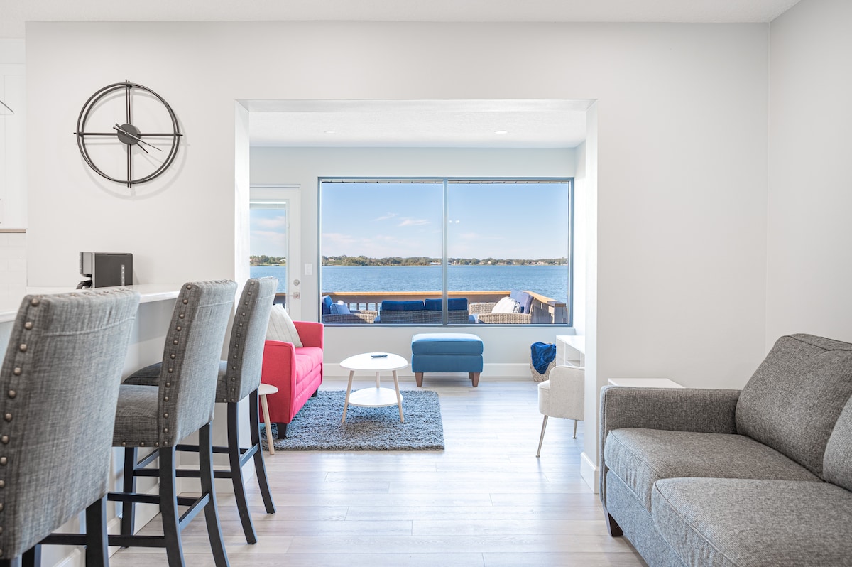 An open living space features large windows framing a scenic view of the lake. Contemporary furniture includes a gray sofa and accent chairs in red and blue. A high-top bar with seating separates the living area from the kitchen, creating a bright and inviting atmosphere.