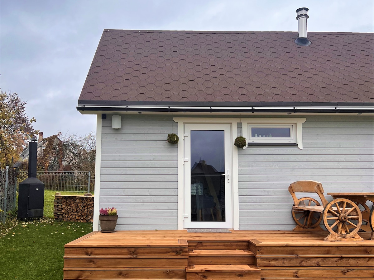 The exterior of the cozy Saunahouse is shown, featuring a wooden deck with a seating area, including a round table and chair. A wood stack is positioned nearby, and a black sauna stove is visible against the grass, creating a serene outdoor environment.
