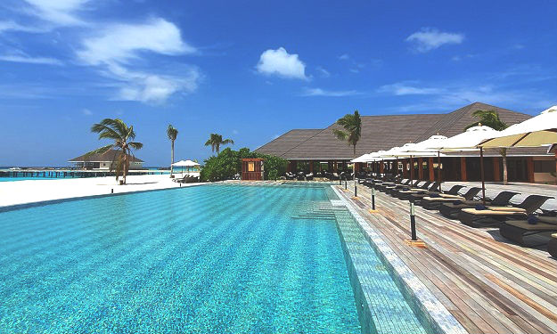 An expansive pool reflects the clear blue sky, surrounded by lounge chairs and umbrellas for shade. The villa's architecture features a thatched roof, blending with lush greenery on one side. The atmosphere suggests a serene outdoor space ideal for relaxation.