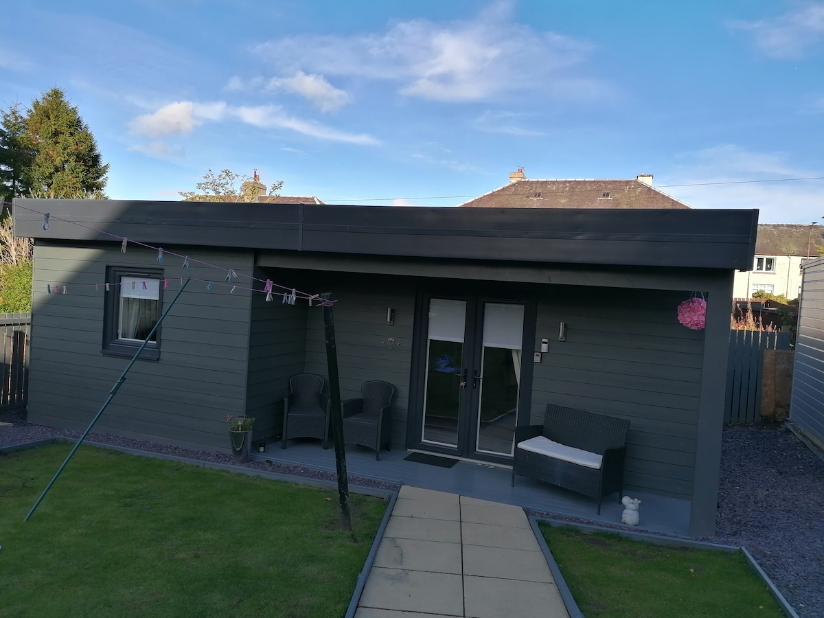 The exterior of Charlotte Cottage is displayed, featuring a modern single-story structure with large glass entry doors. Dark cladding complements the patio area with a light stone pathway. Seating is provided on the front porch, surrounded by a tidy lawn and a clothesline.