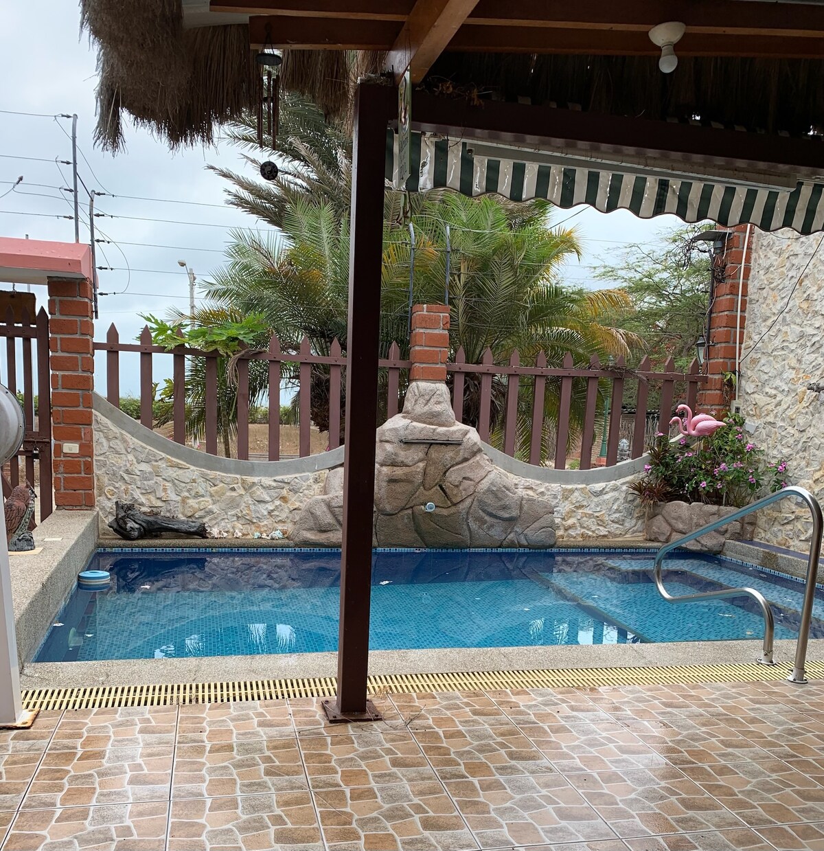 A private pool is surrounded by a stone wall and tropical plants. A shaded area is provided by the roof, and a handrail leads into the water. The pool is in a peaceful setting, accessible for relaxation and leisure.