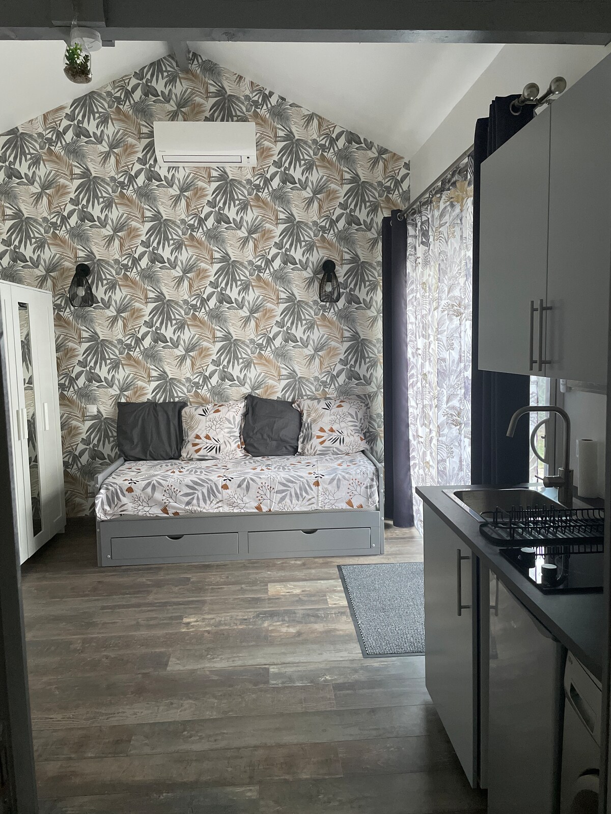 A bright living area features a daybed with patterned cushions against a floral accent wall. The space includes a kitchenette with a gas stove, and natural light enters through a large window dressed with patterned curtains.