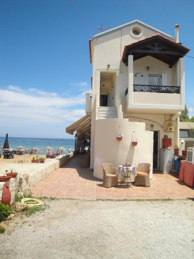 A two-story beachfront apartment is shown, featuring a beige exterior and dark balcony railings. A set of two chairs and a small table is positioned on the patio. The tranquil sea and sandy beach are visible in the background, providing a scenic coastal setting.