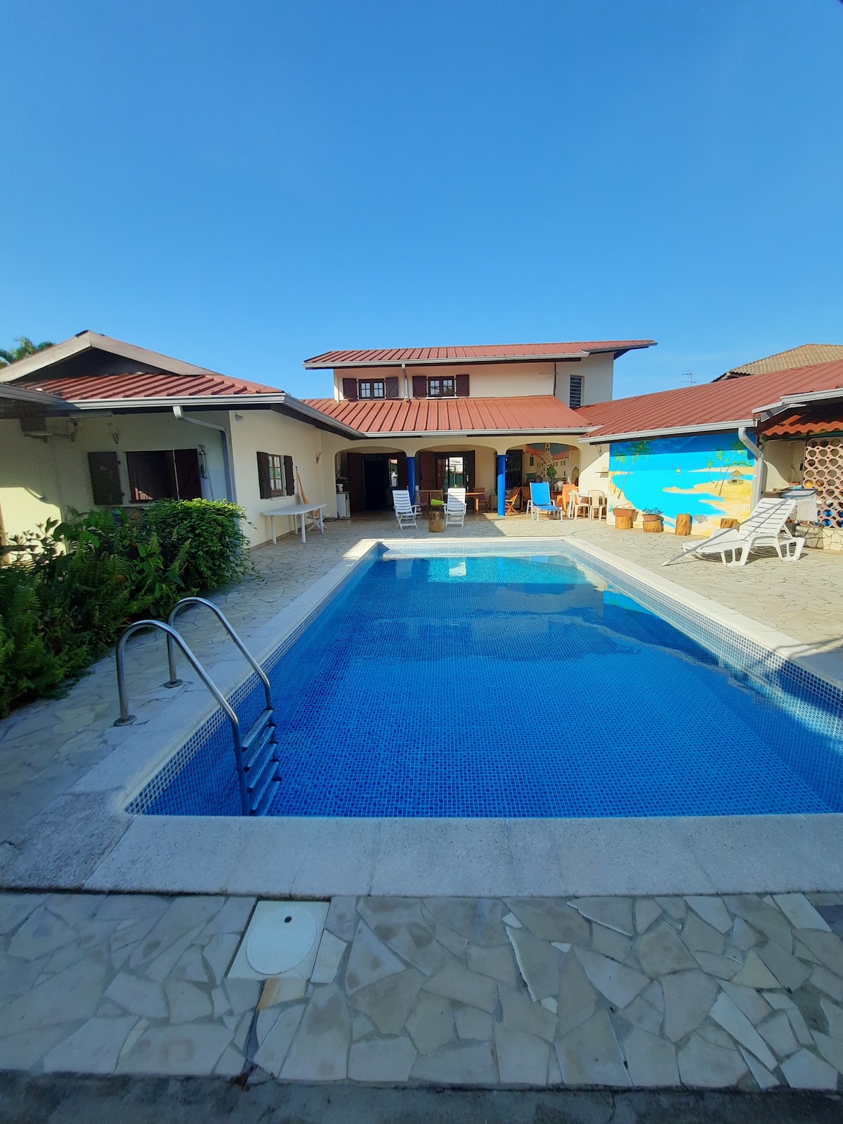A refreshing pool area is featured in this spacious yard, surrounded by sun loungers and greenery. The backdrop showcases a two-story villa with a terracotta roof, and open seating is visible on the patio, creating an inviting space for relaxation.