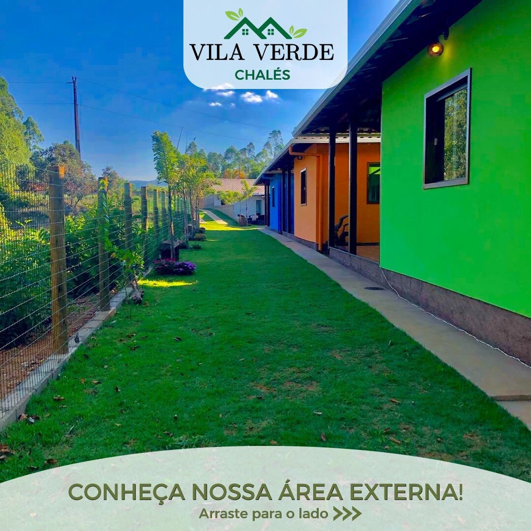 A well-maintained outdoor area is visible, featuring a neatly mowed lawn and colorful chalet exteriors. The path leads alongside the green and blue facades, complemented by trees and landscaping that enhance the natural setting. Fencing encircles the outdoor space, providing privacy.