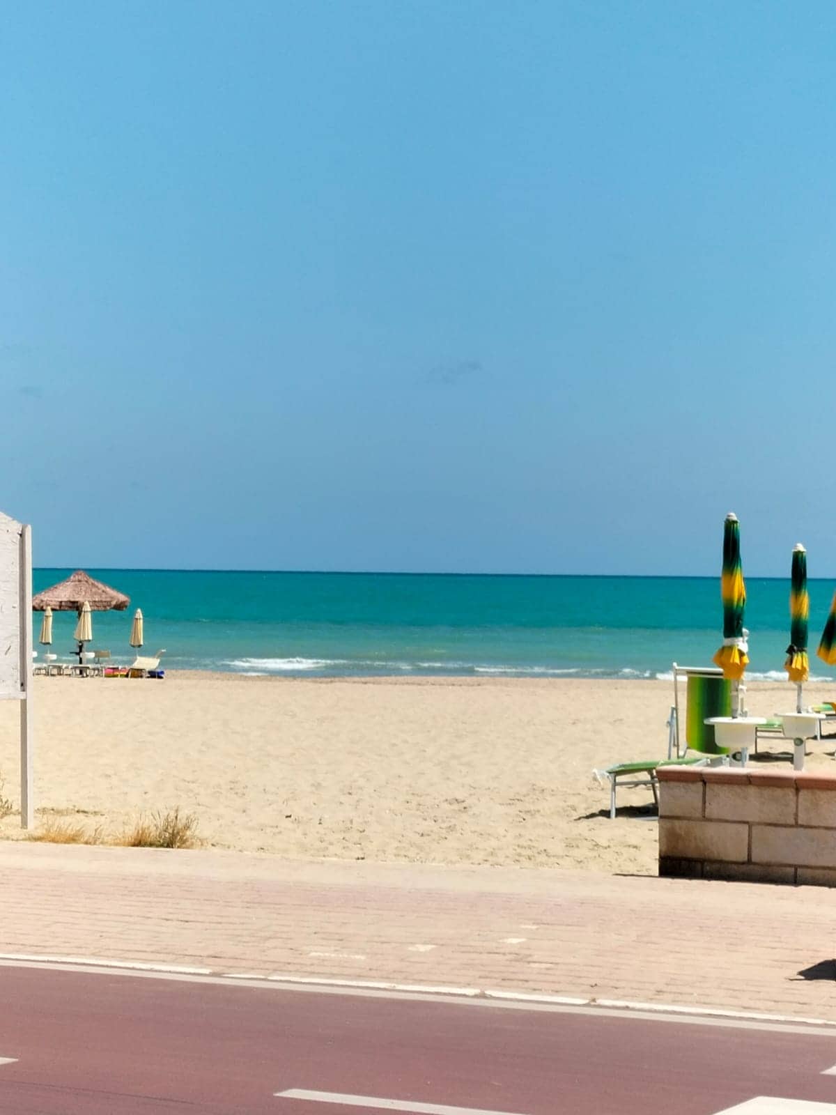 The image showcases a sandy beach with calm turquoise waters extending to the horizon. A thatched-roof gazebo and rows of lounge chairs are positioned on the beach. Colorful umbrella poles stand by the shoreline, providing shade for visitors enjoying the seaside atmosphere.