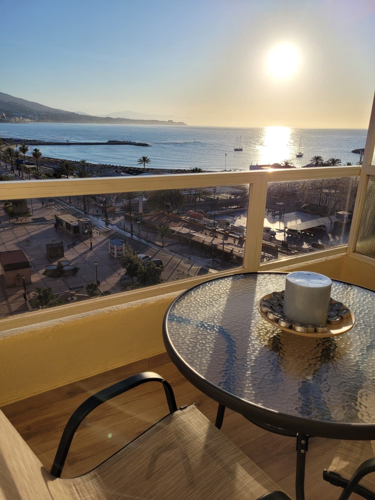 A sunlit balcony offers a clear view of the ocean and distant mountains. A glass table, set with a decorative candle, is accompanied by a single chair. The backdrop features a vibrant sunset reflecting on the water, enhancing the serene coastal atmosphere.