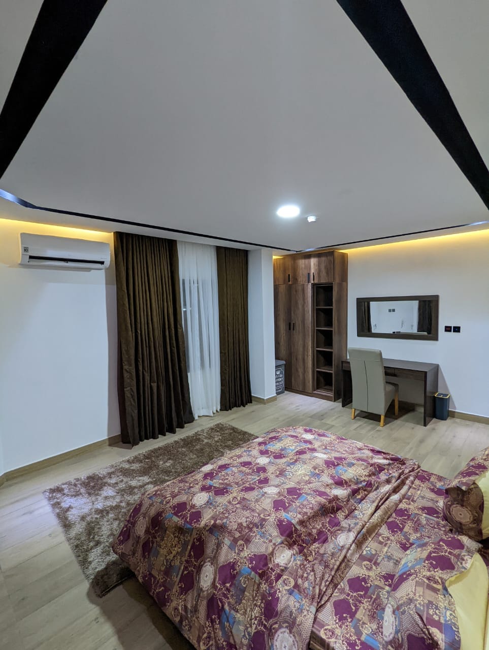 A spacious bedroom features a patterned bedspread draped over a large bed. Dark curtains frame a window, allowing soft light to enter. A wooden desk with a mirror is positioned against the wall, complemented by a comfortable chair. Air conditioning is visible in the corner.