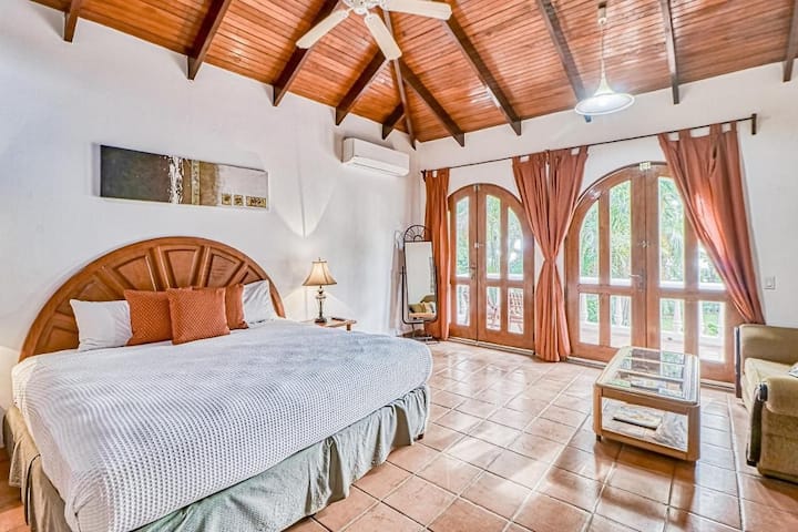 Dormitorio principal muy bien decorado con una gran cama tamaño king y muebles que incluyen mesa de centro y sofá con vistas a través de las dos puertas del arco hacia la amplia terraza/balcón de la casa.
