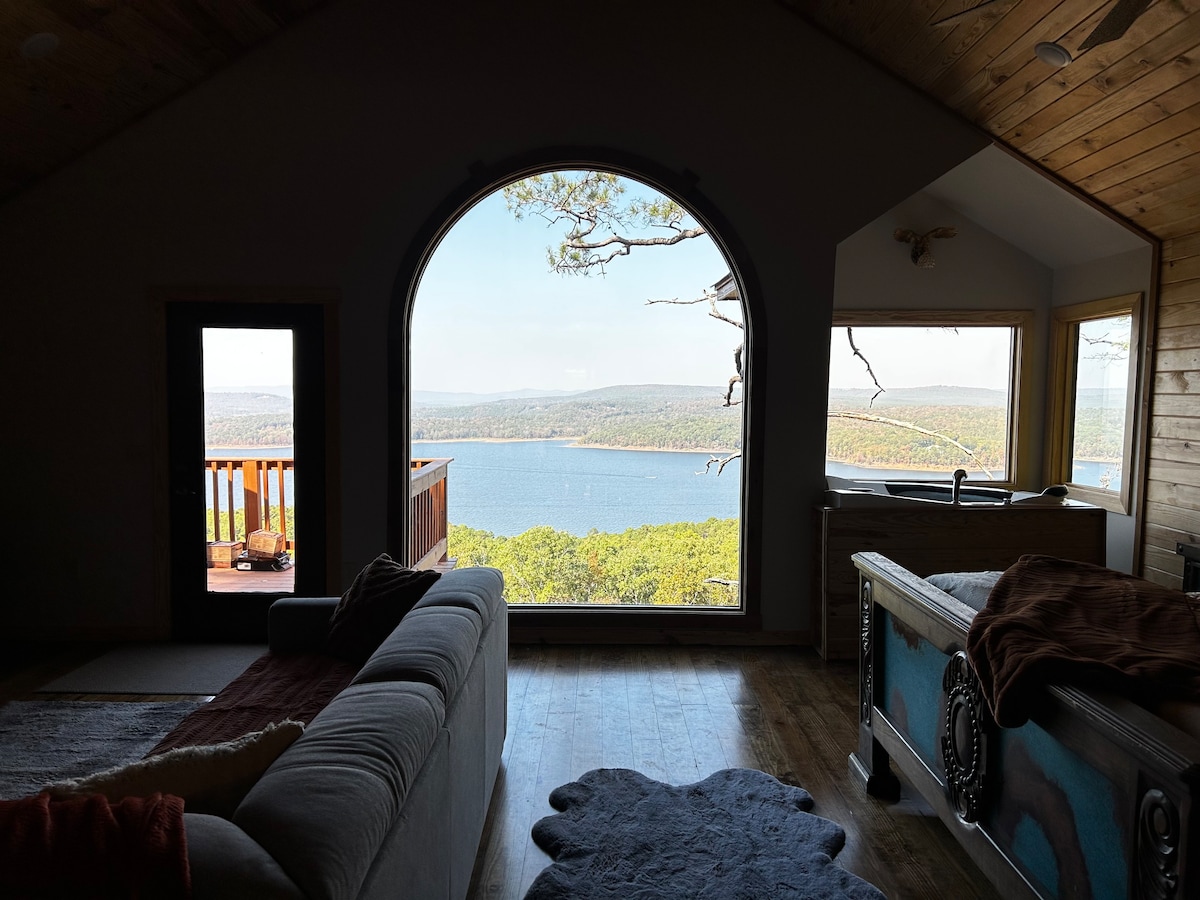 An expansive arched window frames a captivating view of Greers Ferry Lake, with rolling hills in the background. Natural light fills the space, highlighting a spacious sectional couch and a dedicated seating area near the window. A wooden deck is visible outside, enhancing the connection to nature.
