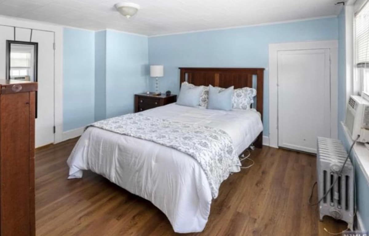 A spacious bedroom features a large bed with light-colored linens and decorative pillows. Blue walls provide a calming backdrop. A window allows natural light to enter, complemented by a bedside lamp. A radiator and an air conditioning unit are present, adding to the room's amenities.