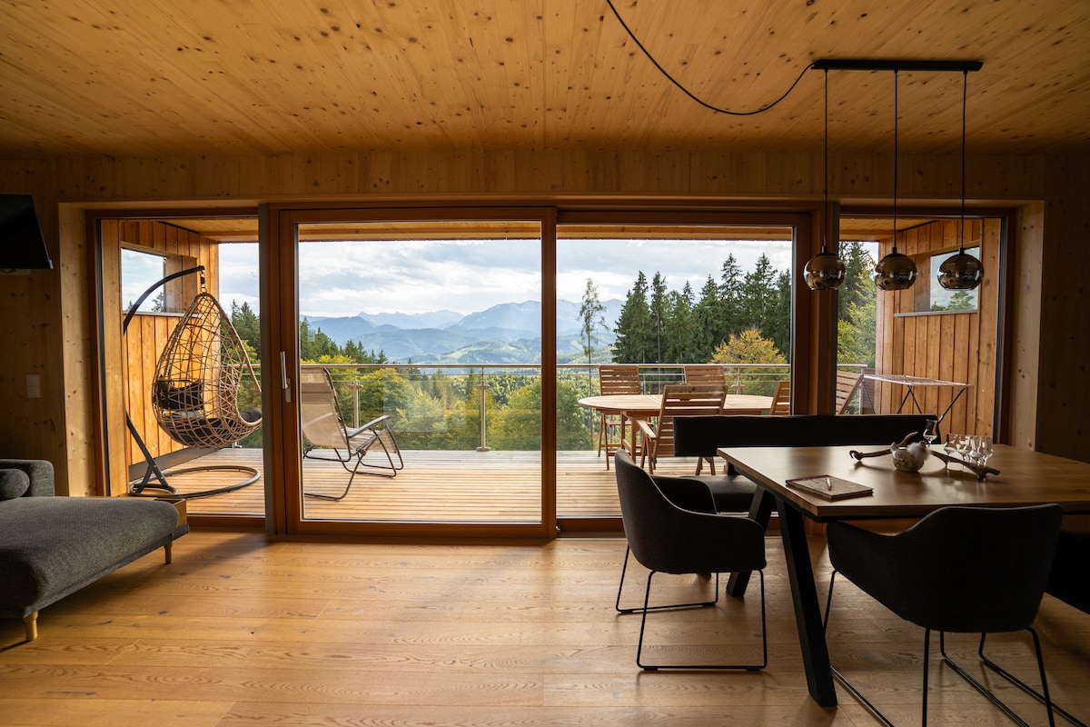 A spacious interior features wooden walls and flooring, enhancing a natural feel. A large sliding glass door opens to a deck with outdoor seating and views of rolling hills and trees. A hammock chair is visible, offering a relaxing spot within the bright living area.