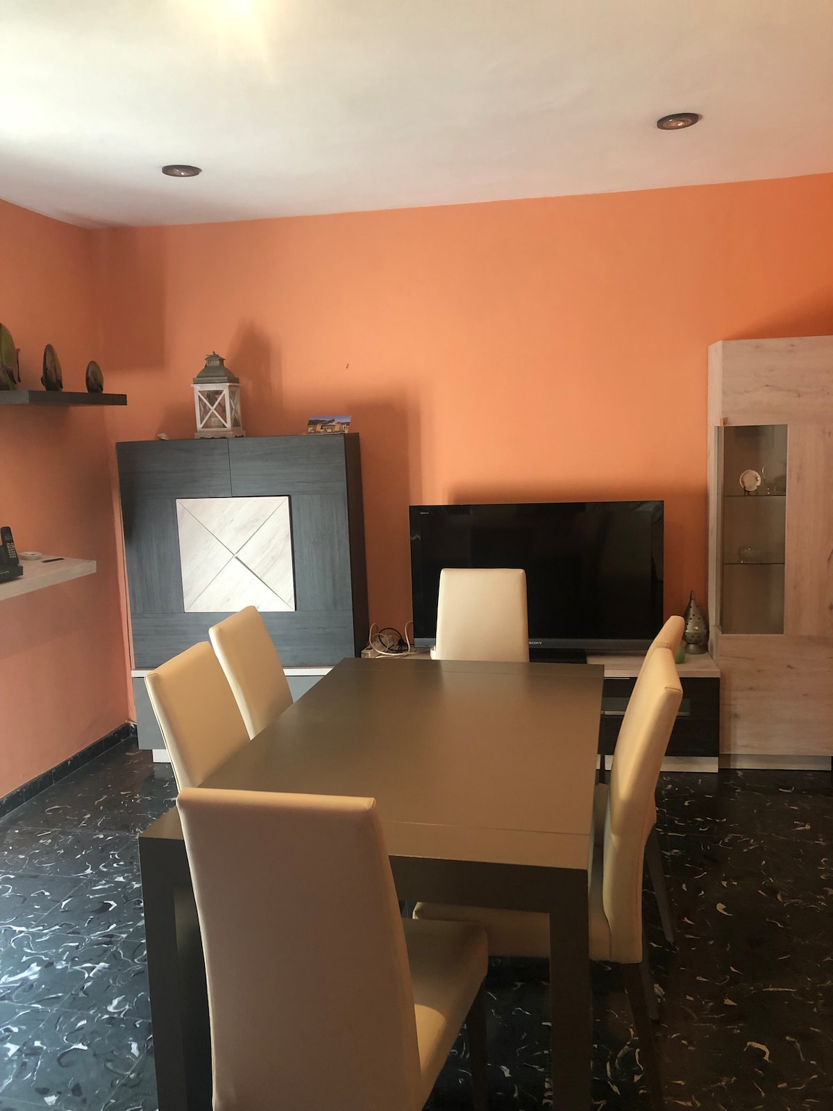 A dining area features a modern table surrounded by four light-colored chairs. An entertainment unit with a television is positioned against an orange wall. Shelves are mounted nearby, adorned with decorative items. The dark floor contrasts with the bright interior.