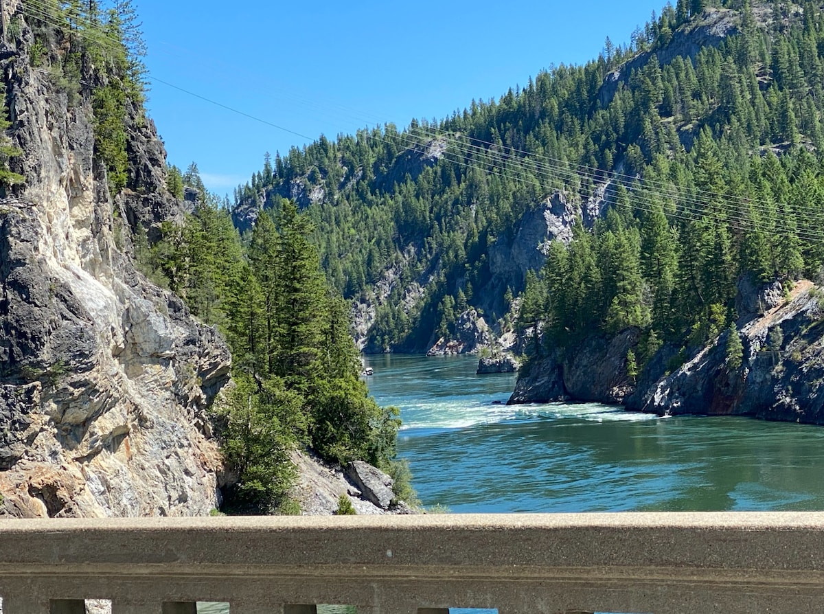 A serene river meanders through the rugged landscape, flanked by steep cliffs and lush greenery. Towering trees line the banks, creating a natural border around the sparkling water. The clear blue sky enhances the tranquil setting, with distant mountains visible in the background.
