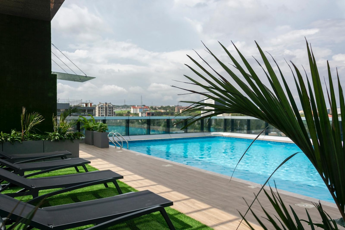A rooftop pool area features a clear blue swimming pool bordered by modern lounge chairs. Lush green plants provide a natural touch along the edge, while expansive views of the cityscape are visible in the background under a partly cloudy sky.
