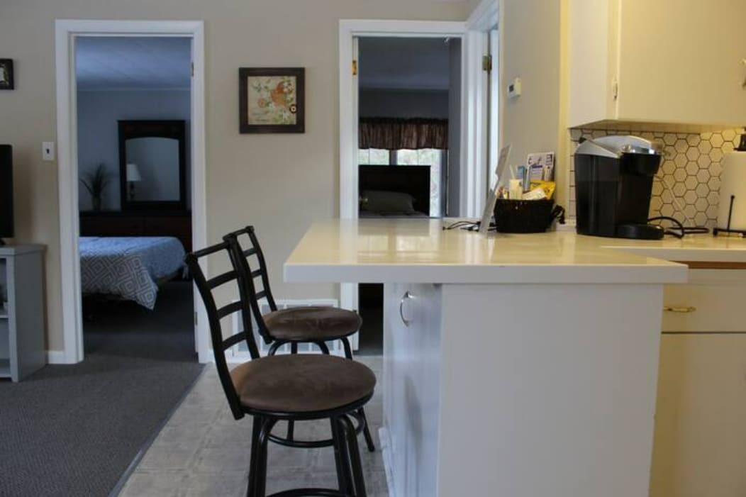 A kitchen area features a countertop with two high chairs. A coffee maker is positioned nearby, and a doorway leads to a bedroom in the background. The neutral wall colors add to the spacious feel of the apartment.