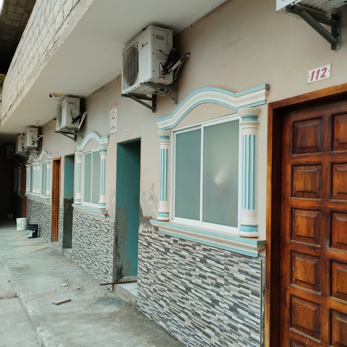 A row of rooms features decorative elements, including arch-shaped window frames and stone accents along the exterior. Each room is equipped with an air conditioning unit above the windows, and a wooden door provides entry. A smooth walkway allows for easy access.