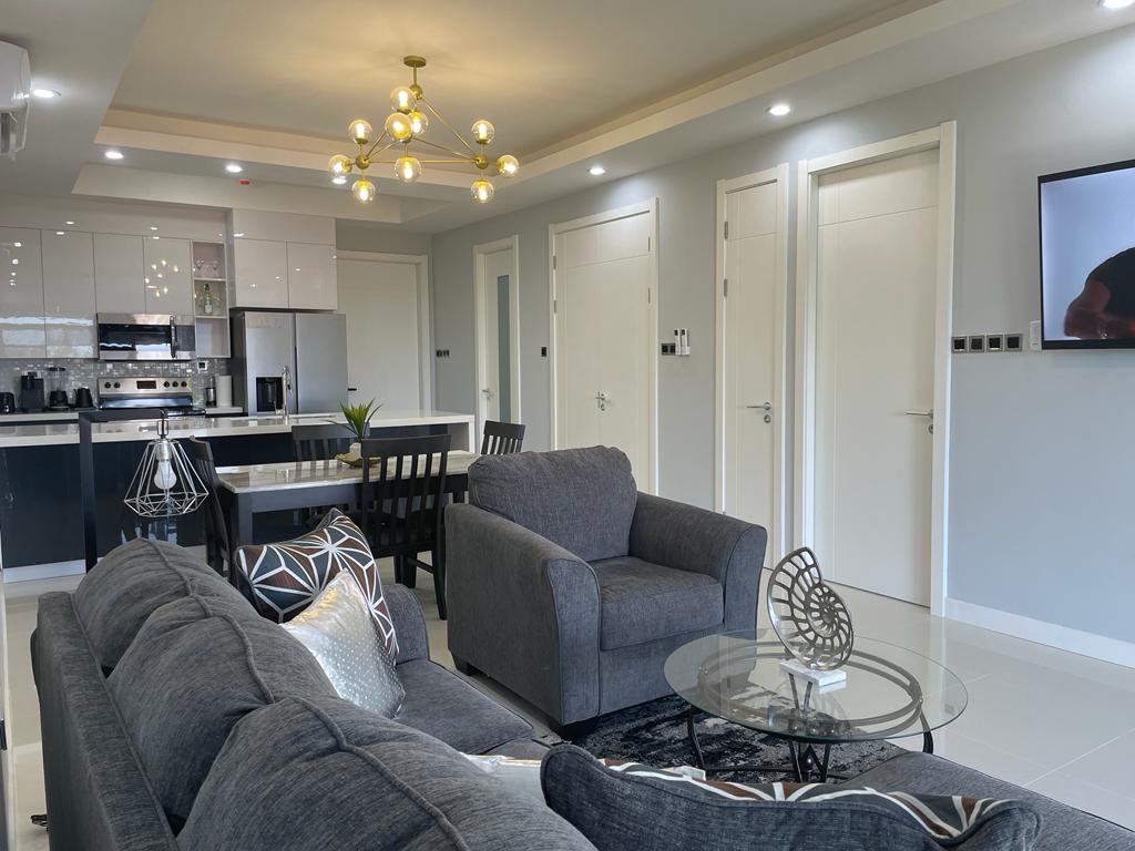 A modern living and dining area features a comfortable gray sofa set and a glass coffee table. A dining table for four is positioned nearby. Contemporary lighting fixtures adorn the ceiling, while appliances are integrated into the sleek kitchen design in the background.