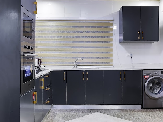 The kitchen features sleek dark cabinetry complemented by golden knobs. A modern stovetop and built-in oven are positioned alongside a spacious countertop. A window with striped blinds allows natural light, and a washing machine is conveniently located within the cabinetry.
