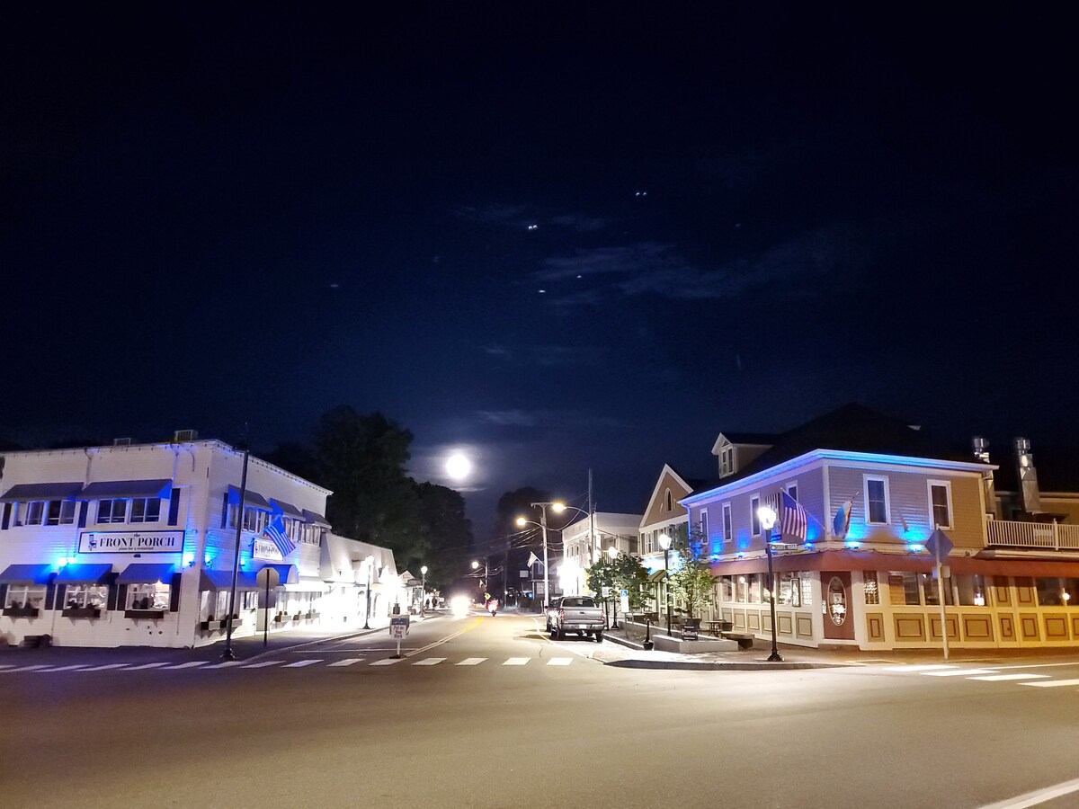 A night scene captures a quiet intersection illuminated by streetlights. Buildings feature decorative blue lighting, with one displaying a sign for a restaurant. A full moon is visible behind wispy clouds in the night sky, adding to the tranquil atmosphere of the downtown area.