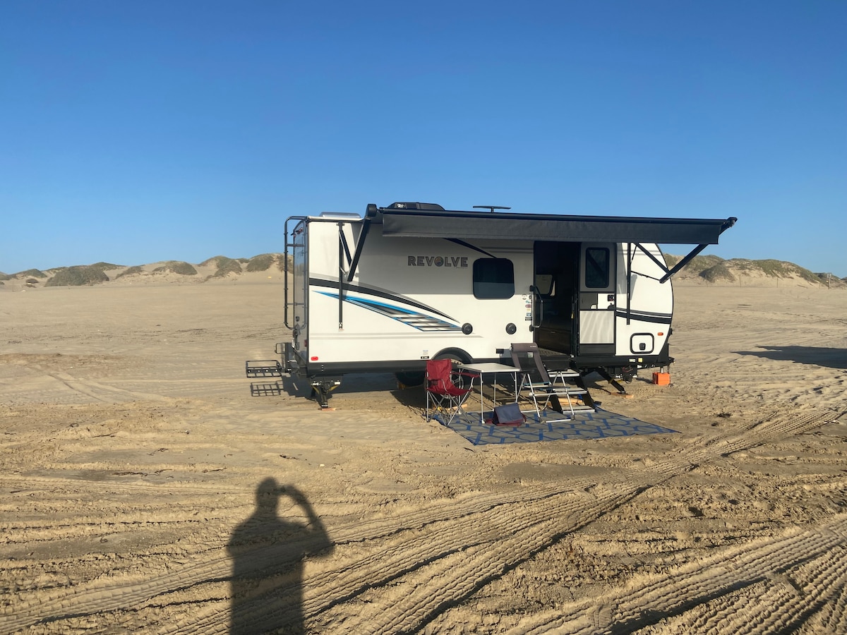 A camper is positioned on a sandy terrain with a vast dune background. An awning extends from the side, providing shade over a seating area with two chairs and a small mat. The sunny sky adds to the outdoor ambiance.