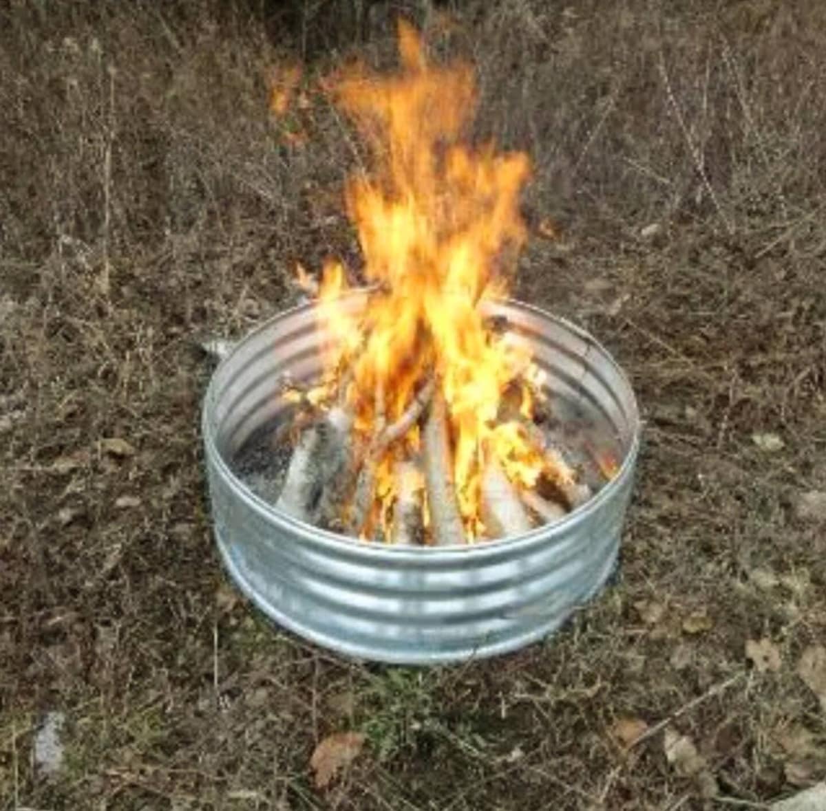 A metal fire ring contains a bright, crackling fire composed of wooden logs. The flames flicker upward, casting a warm glow amid the surrounding dry grass and fallen leaves.