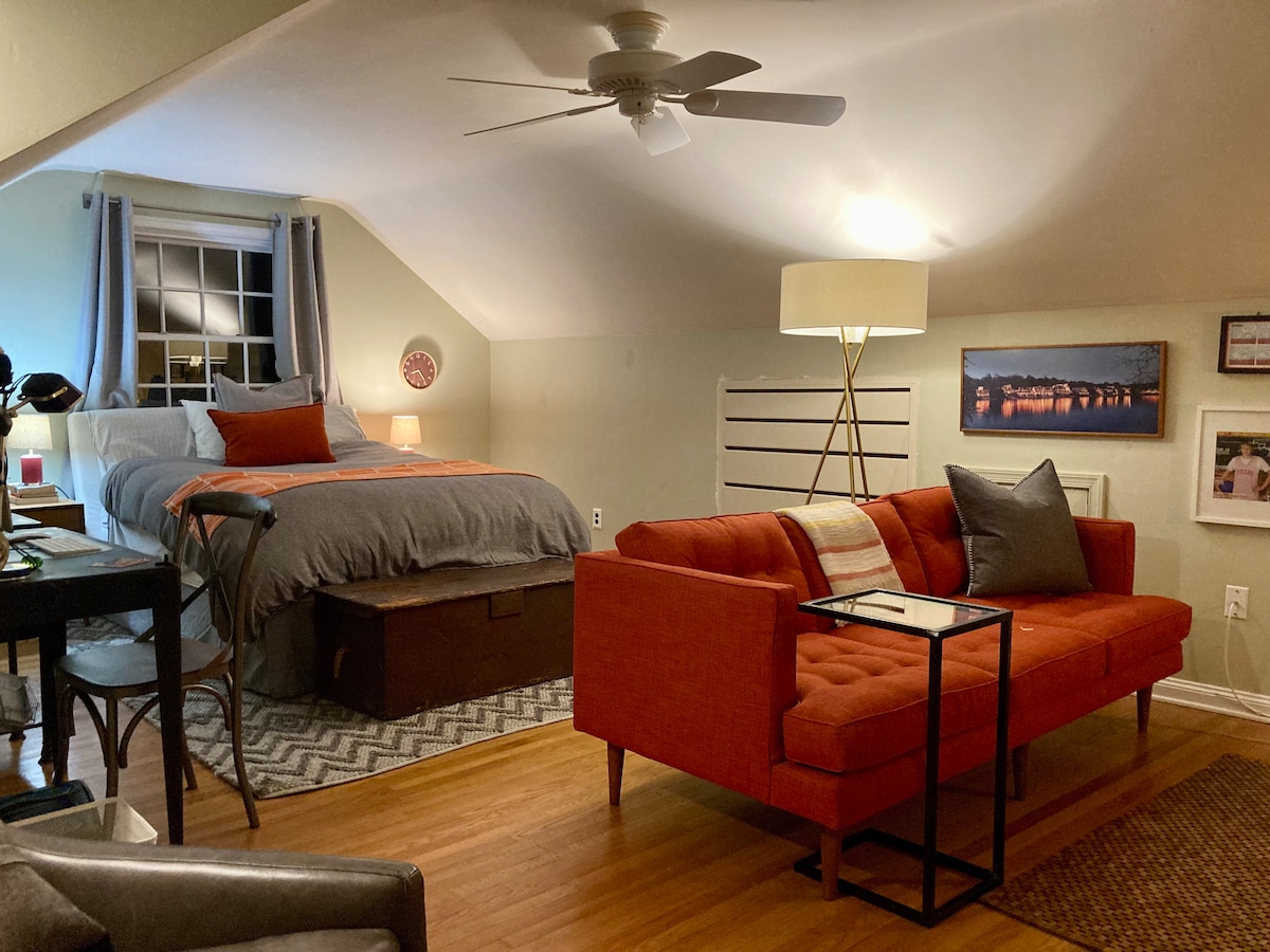 A spacious bedroom is shown with a queen bed dressed in gray linens, accompanied by accent pillows. A cozy orange sofa with decorative cushions sits nearby, alongside a small side table and lamp. Natural light enters through a window, enhancing the inviting atmosphere.