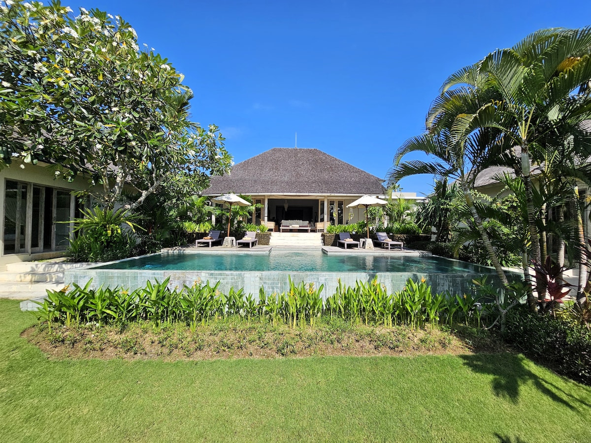A spacious villa is framed by lush greenery and tropical plants, with a tranquil swimming pool in the foreground. Sun loungers and umbrellas are arranged around the pool, offering shaded relaxation. The clear blue sky complements the villa's thatched roof, creating a serene outdoor atmosphere.