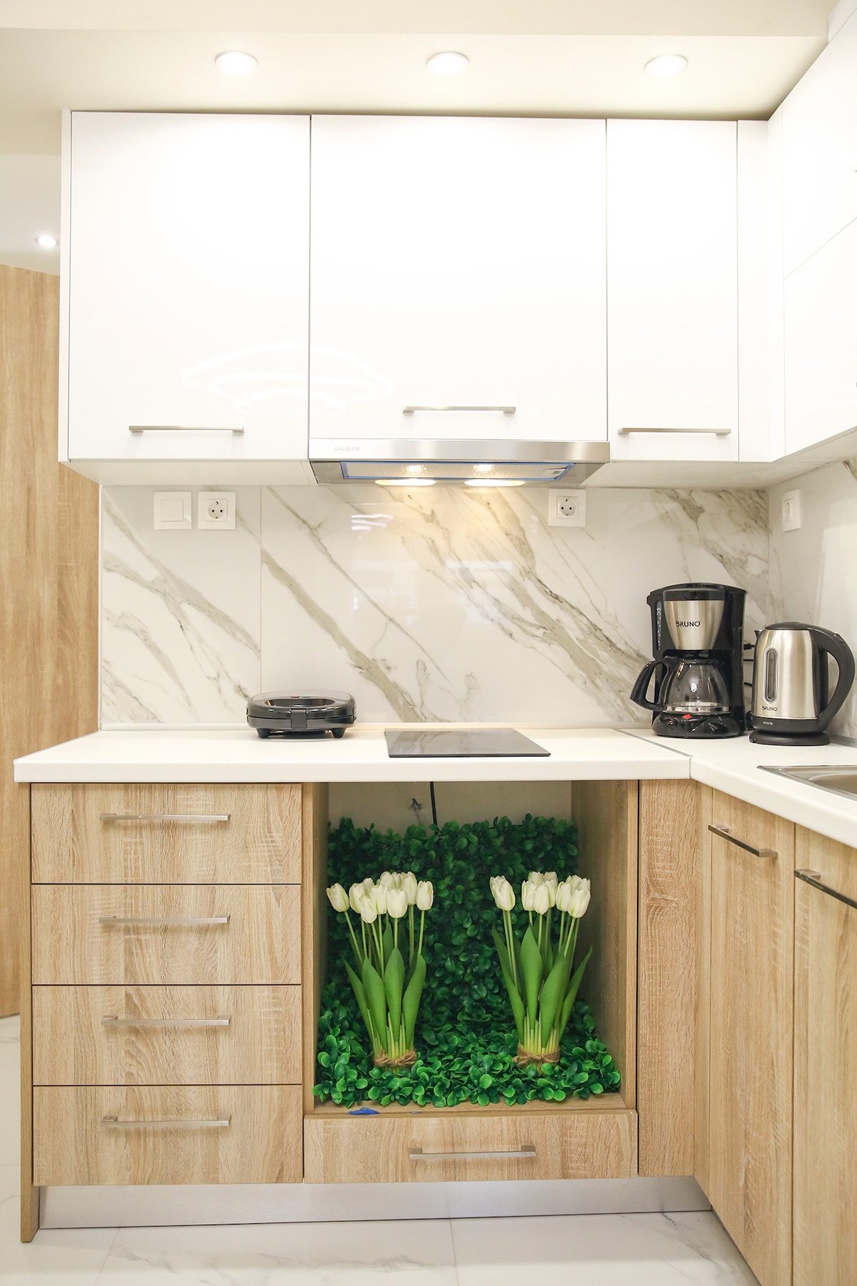 A modern kitchen features sleek white cabinets above a light wood base. A marble backsplash adds elegance, while an organized countertop displays a coffee maker and kettle. Below, a small section is adorned with decorative greenery and white tulips, providing a touch of nature.