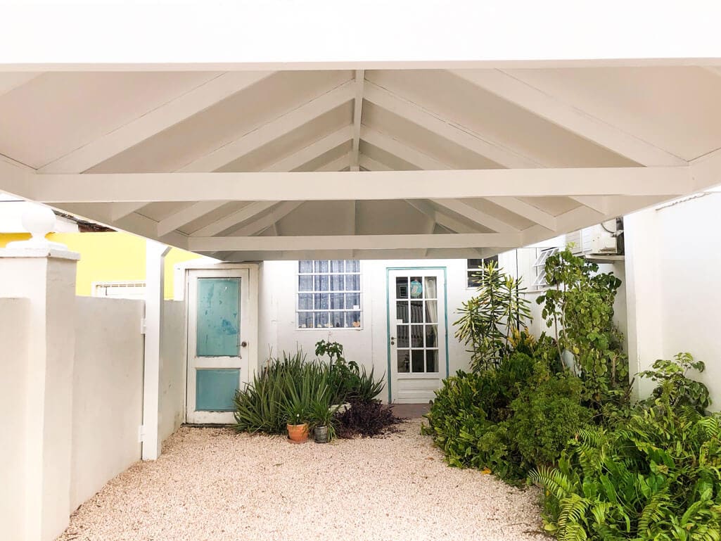 A covered area features a high, slanted roof supported by white beams. The surface is gravel, surrounded by vibrant green plants. Two doors, one painted in soft blue and another in white, lead into the living space. Natural light filters through a nearby window.