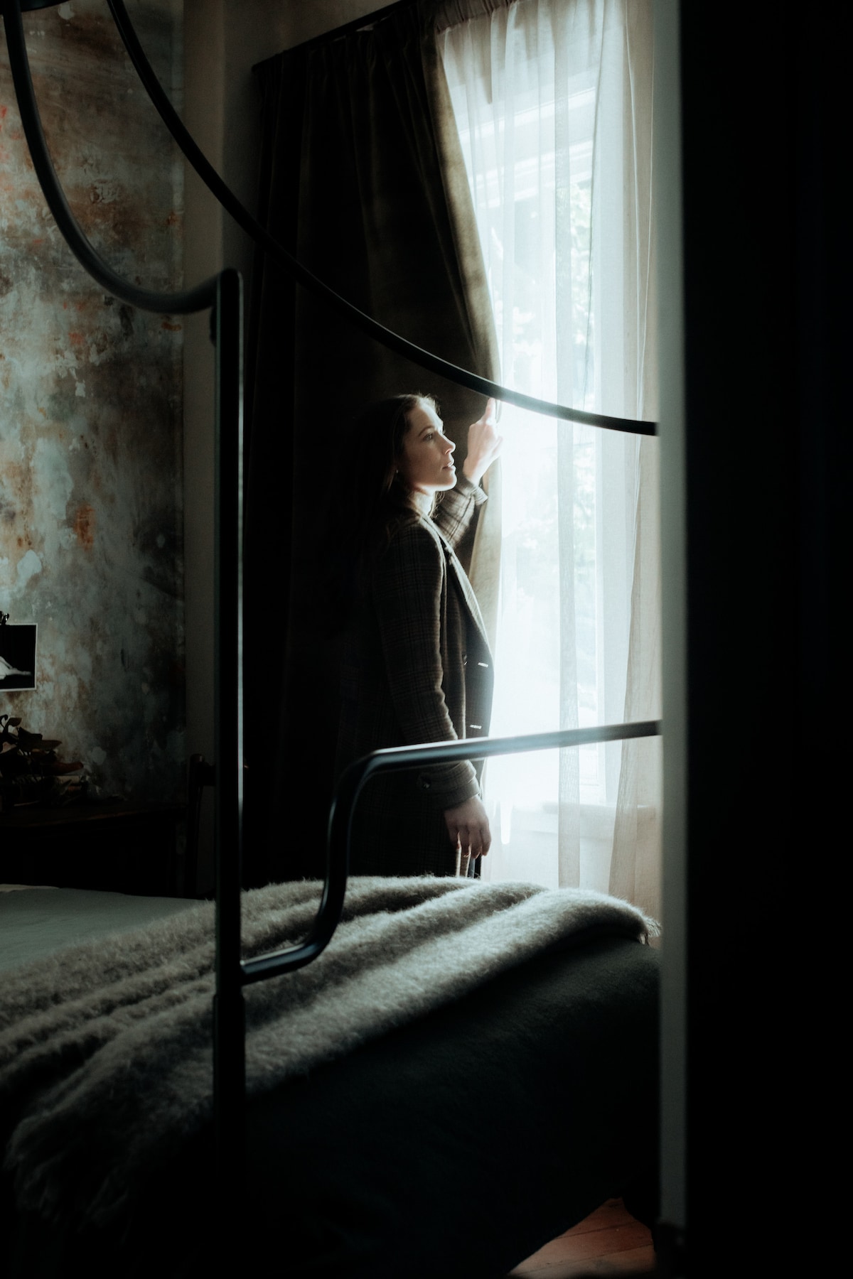 A figure stands by a window, pulling back heavy velvet curtains to allow soft light into the room. In the foreground, a custom-made four-poster bed is partially visible, with textured bedding draping over it.