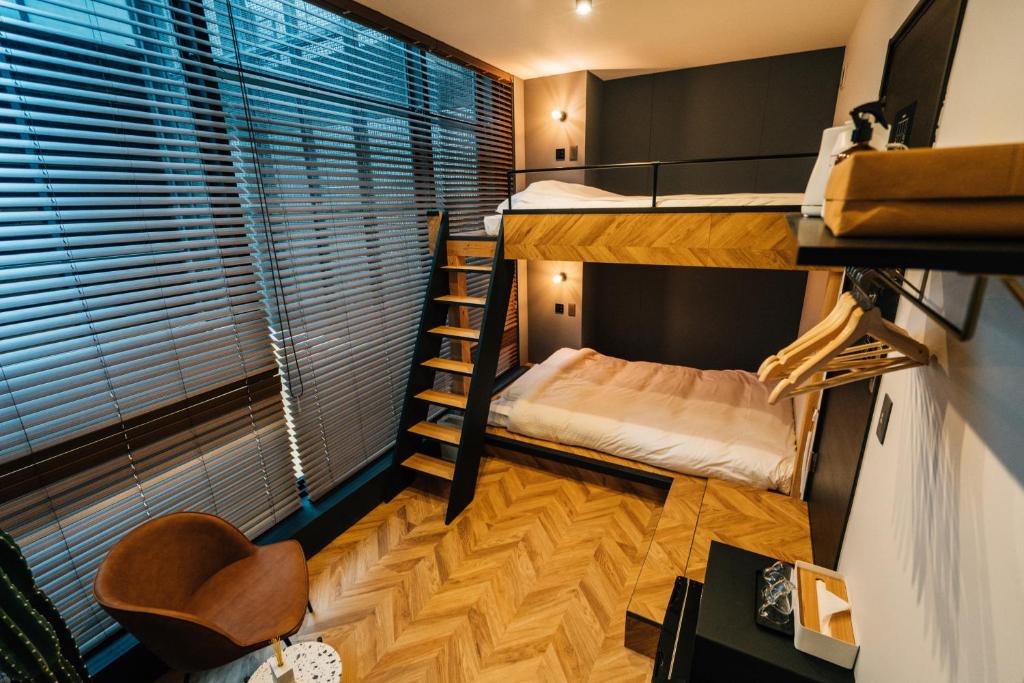 A stylish room features a lofted sleeping area above a spacious bed. Warm wood flooring complements the modern design. A staircase leads to the upper level. Natural light filters through slatted blinds, enhancing the room's cozy ambiance.