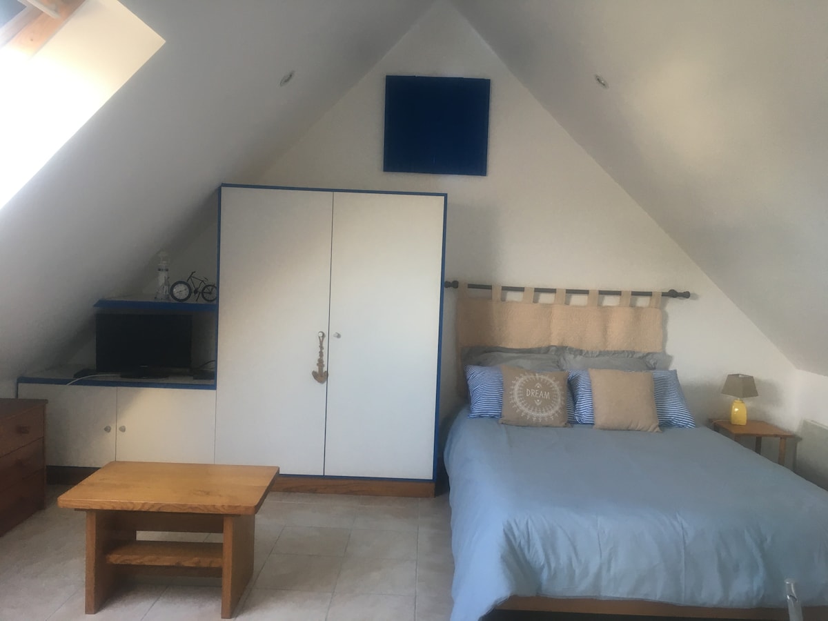 A bright attic room features a spacious bed with soft linens, complemented by decorative pillows. A wooden coffee table stands in front, and a light source is placed beside the bed. The room includes a storage cabinet and a television unit, all set against sloped ceilings.