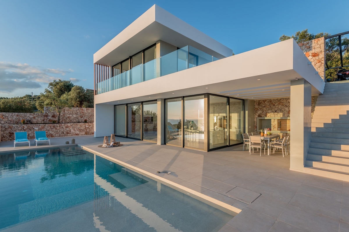 The modern exterior of the villa is prominently displayed with floor-to-ceiling glass walls reflecting the tranquil pool. The private pool area includes lounge chairs, while an outdoor dining space is visible under a shaded terrace, offering serene views of the surrounding landscape.