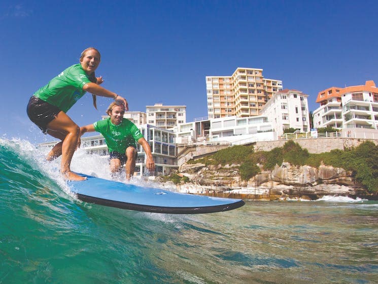 Two individuals are depicted surfing on a wave, showcasing the dynamic activity of the sport. A shoreline of multi-story buildings serves as a backdrop, with lush greenery visible along the rocky coastline. The clear blue sky completes the vibrant beach scene.