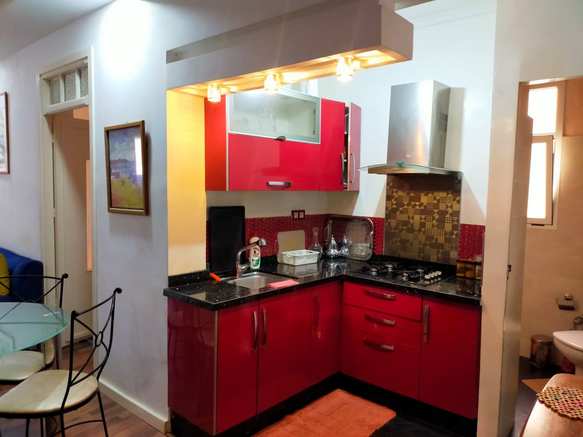 A modern kitchen features vibrant red cabinets and black countertops. Stainless steel appliances include a gas stove and a chimney. The dining area is visible with a glass table and iron chairs, while natural light enters through a nearby window.
