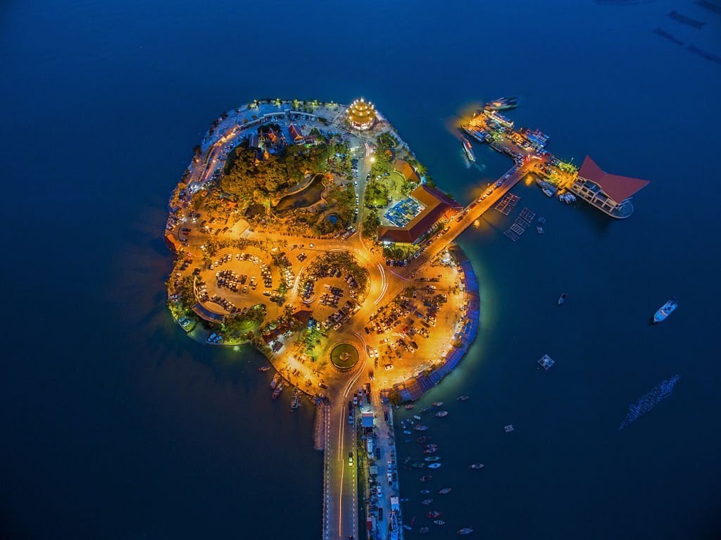 An aerial view captures a vibrant island illuminated by warm lights against the dark water. The layout of roads and pathways winds through lush greenery and areas of activity. A dock is visible, accommodating boats, while the surrounding sea reflects the glimmering lights.