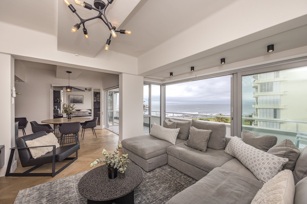 A spacious living area features a sectional sofa adorned with neutral-toned cushions, oriented towards large floor-to-ceiling windows that provide views of the ocean. A circular coffee table with a floral arrangement sits center stage, while a dining area with a round table is visible in the background.