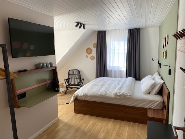 A cozy room features a spacious bed with white linens against a light green accent wall. A flat-screen TV is mounted above a minimalist shelf, and a comfortable chair is positioned near a window covered with sheer curtains. Natural light enhances the wooden floor.