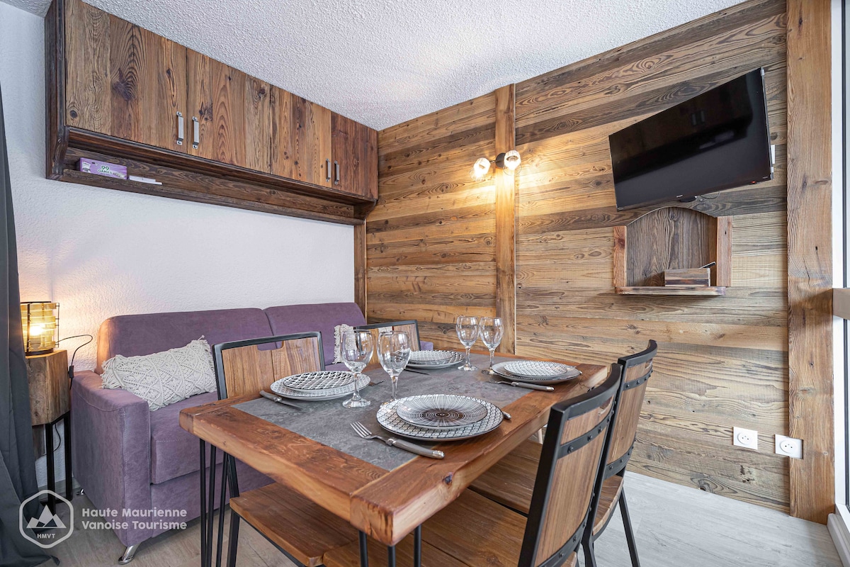 A cozy living area is featured, showcasing a wooden dining table set for four, accompanied by black chairs. A purple sofa provides seating, and a mounted TV is visible on the wall. Natural wood paneling adds warmth to the space.