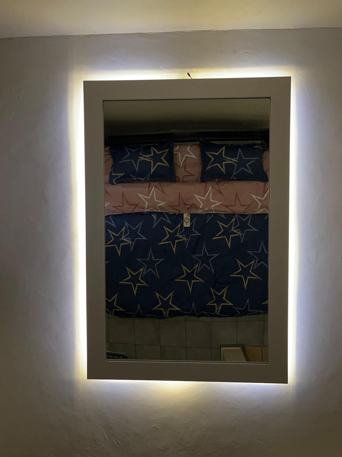 A rectangular mirror is framed with a soft glow of LED lighting. Reflected in the mirror is a bed adorned with a duvet cover featuring a star pattern in blue and gold, with matching pillows. The overall ambiance conveys a calm and cozy sleeping space.