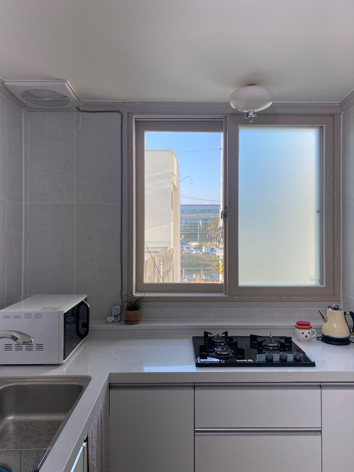 A bright kitchen space features a large window allowing natural light. A gas stove with multiple burners is positioned beside a sink. Nearby, a microwave sits on the countertop, accompanied by a small kettle and a decorative item.